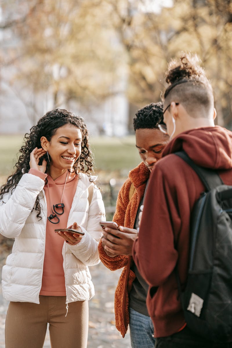 Diverse friends in casual outfits standing in fall park and surfing mobile phones while spending time together